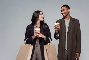 happy interracial couple in stylish outfits holding shopping bags and paper cups isolated on grey
