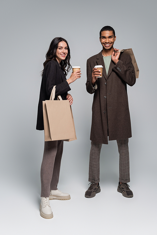 full length of cheerful interracial couple in stylish outfits holding shopping bags and paper cups on grey