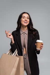 happy young woman holding paper cup with coffee to go and shopping bags isolated on grey