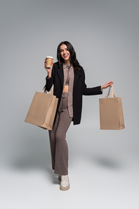 full length of cheerful young woman holding paper cup with coffee to go and shopping bags on grey