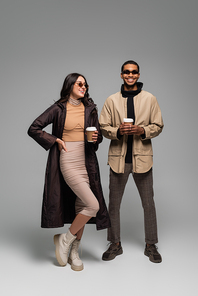 full length of cheerful interracial couple in stylish outfits and sunglasses holding paper cups on grey