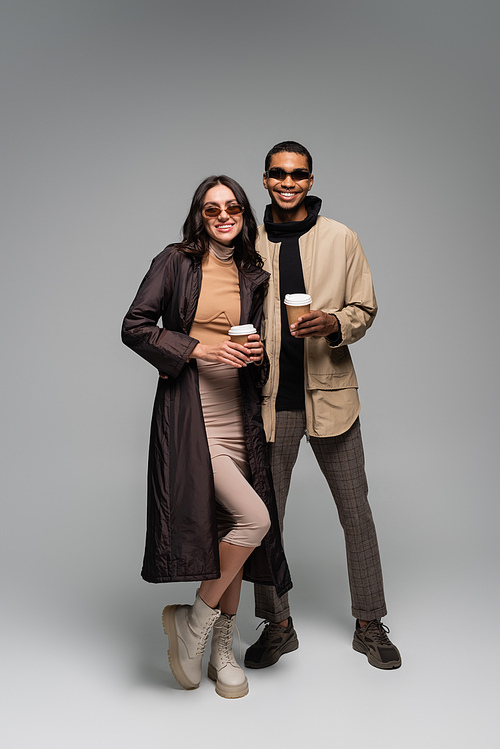 full length of happy interracial couple in stylish outfits and sunglasses holding paper cups on grey