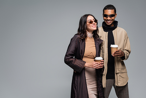 cheerful interracial couple in stylish outfits and sunglasses holding paper cups isolated on grey