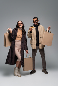 full length of happy interracial models in stylish sunglasses holding shopping bags and credit card on grey