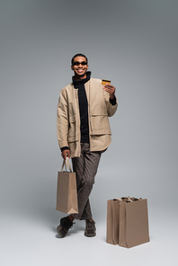 full length of happy african american man in sunglasses and autumnal outfit holding credit card and shopping bag on grey