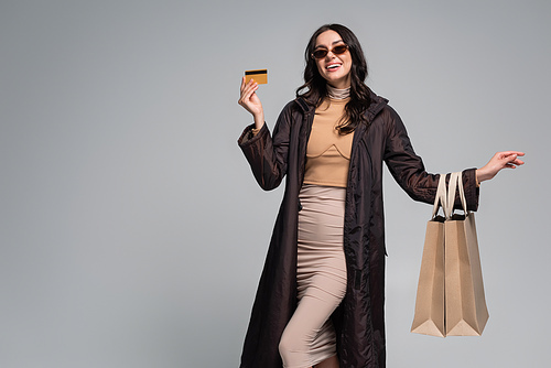 smiling young woman in stylish sunglasses holding shopping bags and credit card isolated on grey
