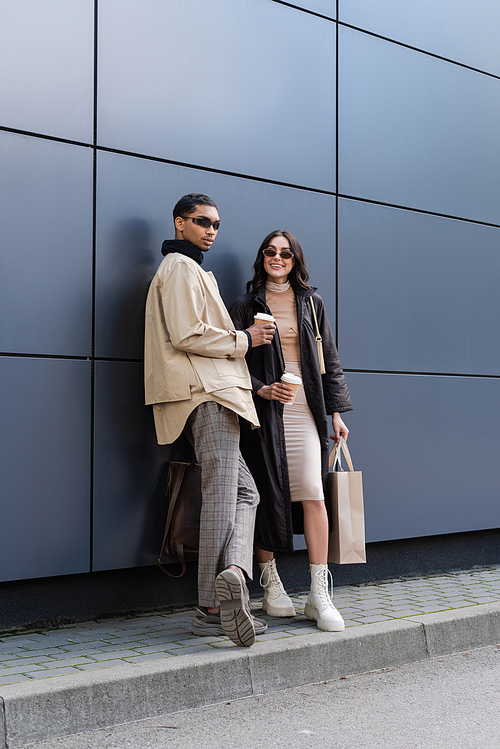 stylish african american man and happy young woman in sunglasses holding paper cups and shopping bag