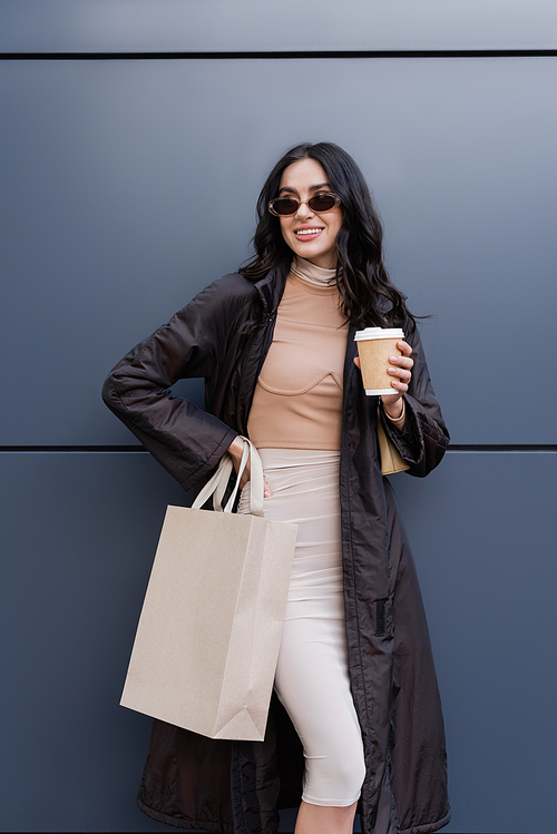 happy young woman in stylish outfit and sunglasses holding paper cup and shopping bag near building