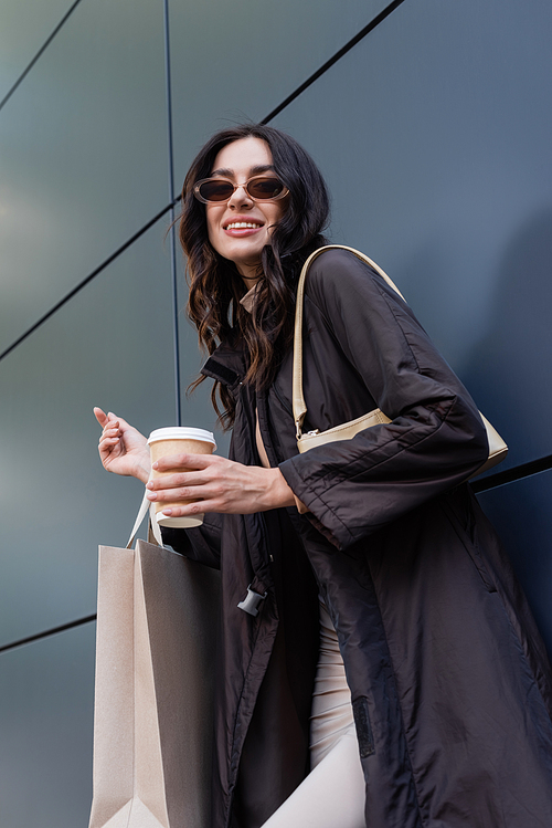 happy brunette woman in stylish outfit and sunglasses holding paper cup and shopping bag near building