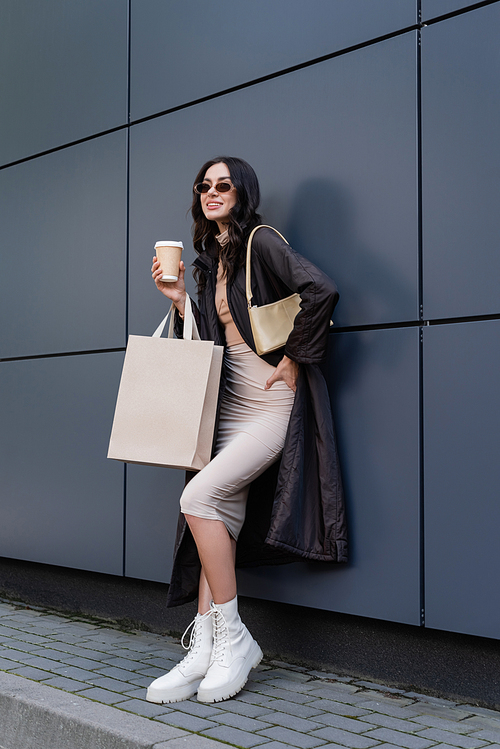 brunette woman in stylish outfit and sunglasses holding paper cup and shopping bag while smiling near building