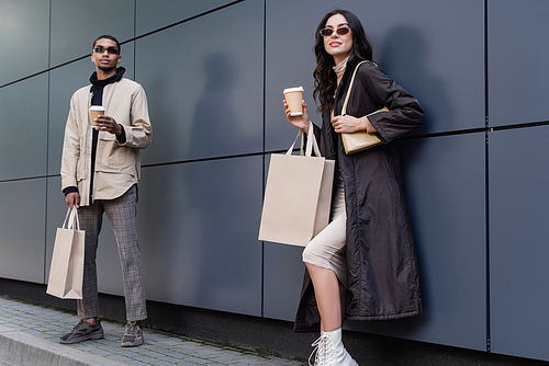 young stylish woman with handbag and paper cup holding shopping bag near african american man