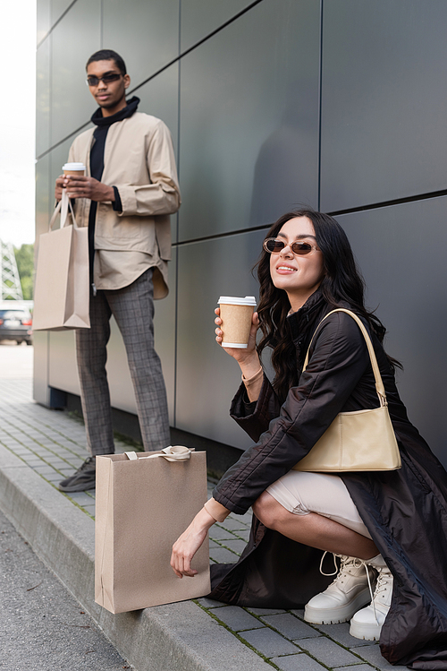 young stylish woman with handbag and paper cup near shopping bag and blurred african american man