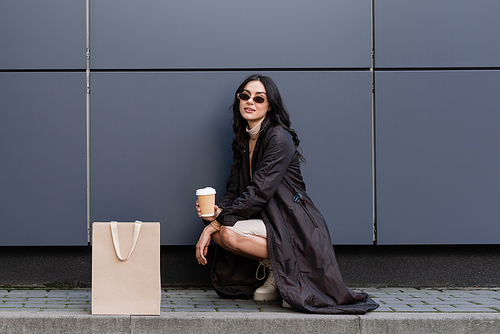 young stylish woman with paper cup sitting near paper bag and building of shopping mall