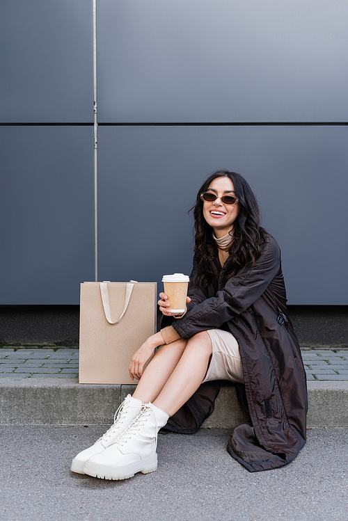 young happy woman with paper cup sitting near paper bag and building of shopping mall