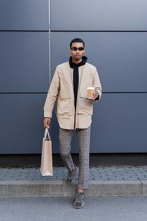 full length of stylish african american man in sunglasses and autumnal outfit holding paper cup and shopping bag