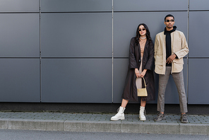 full length of young multiethnic couple in trendy autumnal outfits and sunglasses posing near building