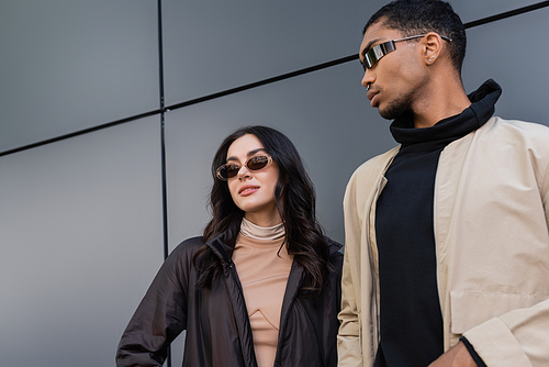 low angle view of stylish multiethnic couple in trendy autumnal outfits and sunglasses standing near building