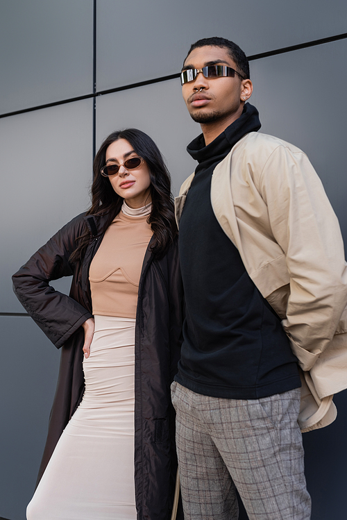 stylish multiethnic couple in autumnal outfits and trendy sunglasses standing near building