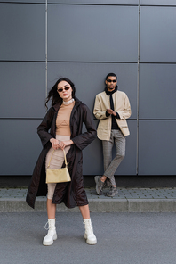 full length of stylish young woman in autumnal coat and sunglasses standing with handbag near african american man