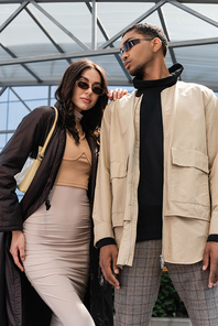 low angle view of stylish interracial couple in sunglasses and stylish outfits looking at camera