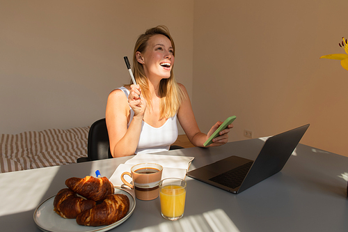 Excited freelancer holding smartphone and pen near laptop and breakfast at home