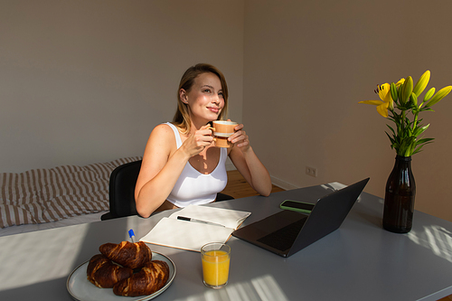 Pleased freelancer holding cup of coffee near gadgets and croissants at home