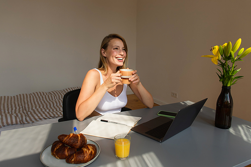 Happy blonde woman holding cup near breakfast and gadgets at home