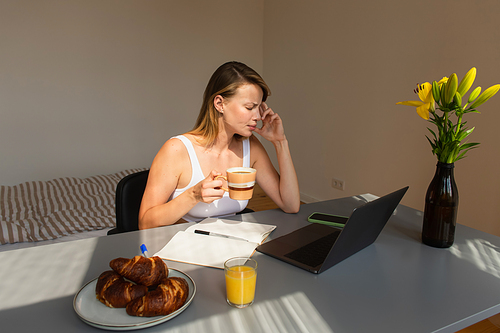 Side view of freelancer holding cup while suffering from headache near devices and breakfast at home