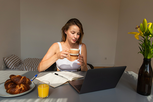 Blonde woman holding cup and looking at laptop near flowers and breakfast at home
