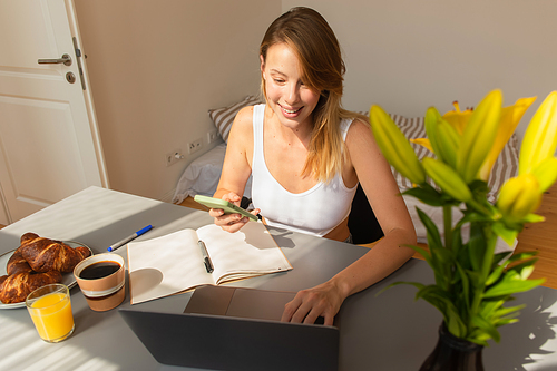 Smiling woman using devices near notebook and breakfast at home