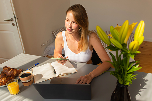 Blonde freelancer using smartphone and laptop near breakfast at home