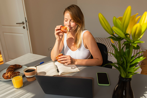 Freelancer holding croissant near devices and drinks at home