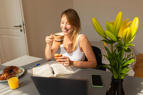 Cheerful freelancer holding coffee and croissant near gadgets and flowers at home