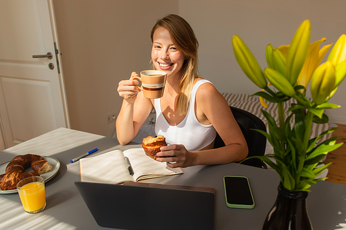 Positive freelancer holding coffee and croissant near devices and flowers at home