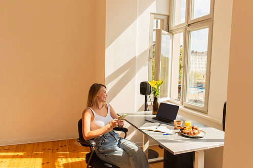 Side view of smiling freelancer using smartphone near laptop and breakfast at home