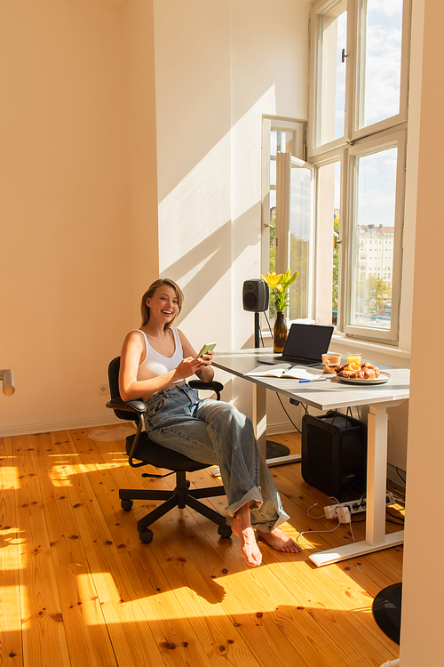 Positive woman using smartphone near laptop and breakfast at home