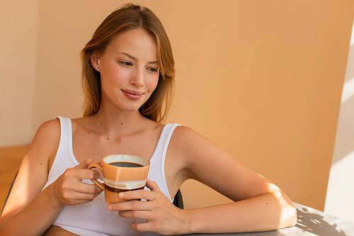 Blonde woman in top holding cup of coffee at home in morning