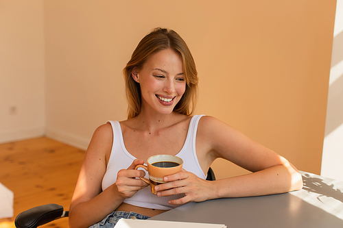 Positive blonde woman holding cup of coffee near notebook at home