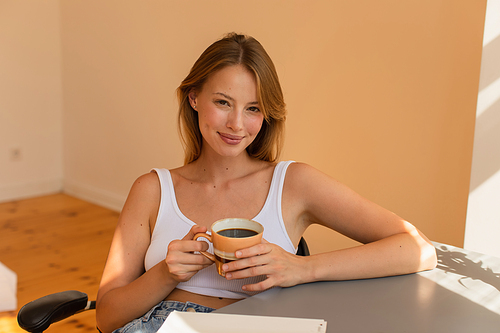 Blonde woman in top holding cup of coffee and looking at camera near notebook at home
