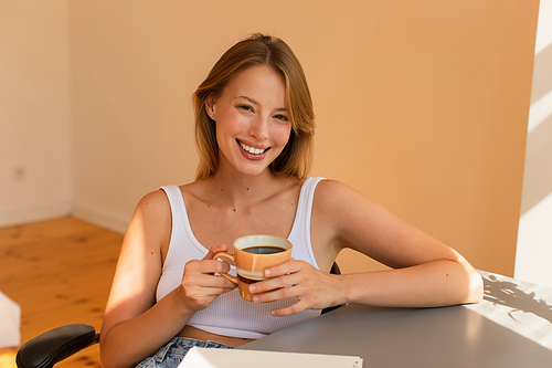 Cheerful blonde woman holding cup of coffee near notebook on table at home