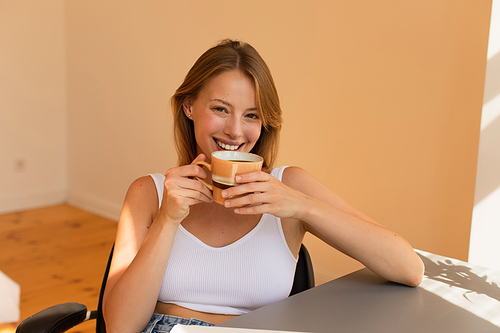 Happy blonde woman in top holding cup of coffee at home
