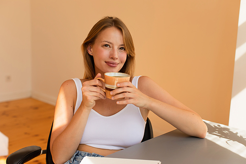 Dreamy blonde woman in top holding cup of coffee at home