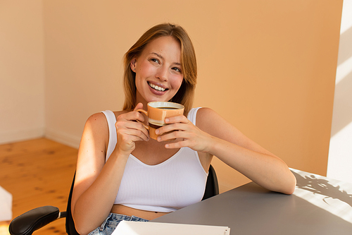 Cheerful young woman in top holding cup of coffee near table at home