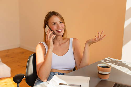 Smiling blonde woman talking on smartphone near coffee and laptop at home