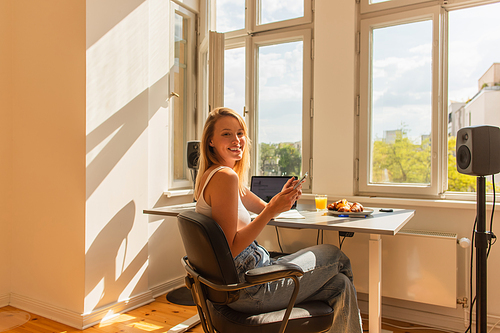 Happy blonde freelancer using cellphone near orange juice and breakfast at home