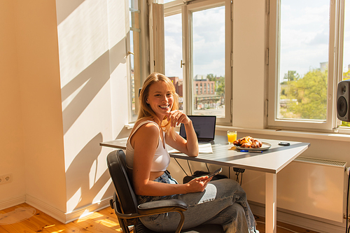 Positive blonde freelancer looking at camera near devices and breakfast at home