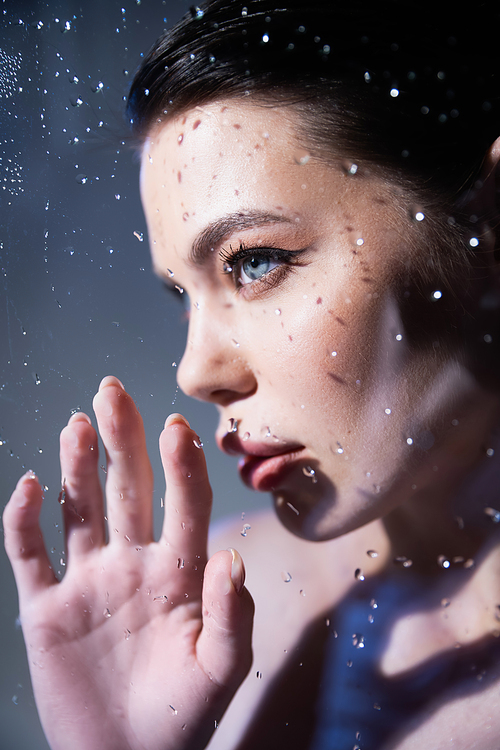 Young model with makeup touching glass with water drops on grey background