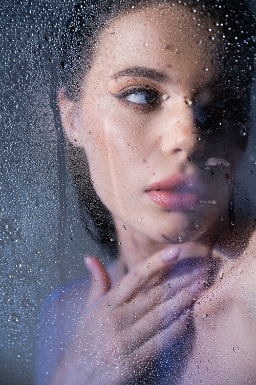 Young brunette woman touching neck behind wet glass on grey background