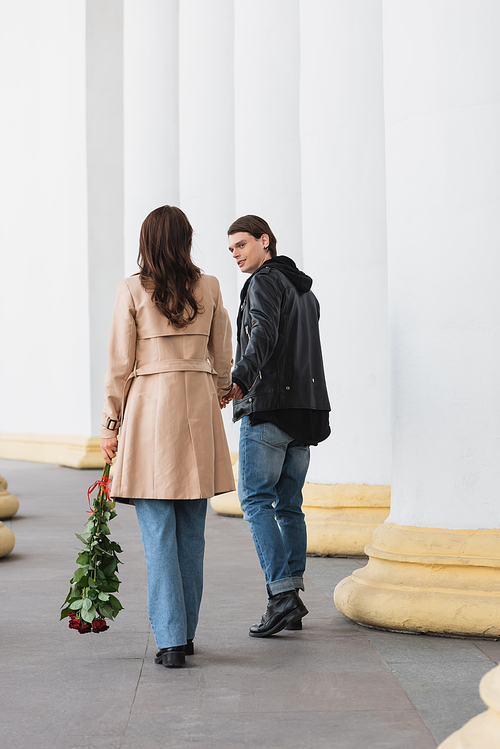 full length of happy young man holding hand of girlfriend in trench coat standing with red roses