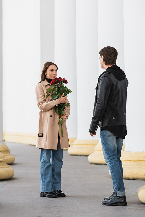 full length of pretty young woman holding red roses and looking at stylish boyfriend in jacket